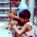 A boy beats on a drum while his father plays a Thai saw.